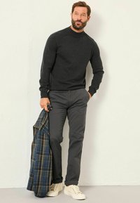 Dunkelgrauer Strickpullover mit hohem Kragen, kombiniert mit grauen Hosen und weißen Sneakers. Hält eine blaue Karojacke.