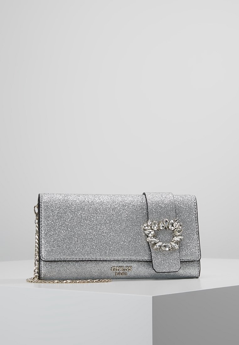 Pochette argentée pailletée avec boucle ornée de cristaux et bandoulière en chaîne dorée, présentée sur un piédestal géométrique blanc.