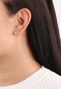 Gold-tone flower-shaped stud earrings with clear gem accents, featuring leaf details, worn on a model's earlobe against dark hair.