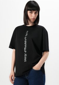 Juoda per didelė medvilninė marškinėlių su trumpomis rankovėmis, su vertikaliu baltu logotipo grafika "KARL LAGERFELD JEANS" priekyje. Laisvas siluetas.