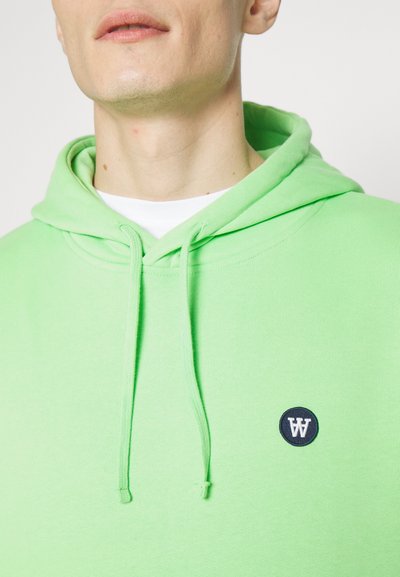 Grønn bomullhettetrøye med snøring, ribbestrikkede mansjetter og et sirkulært marineblått logo-merke på brystet. Glatt tekstur, avslappet passform.