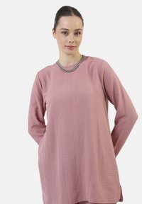 Maglia a maniche lunghe in rosa polveroso, con una texture stropicciata, scollo rotondo e spacchi laterali per facilitare i movimenti. Adornata con una catena argentata.