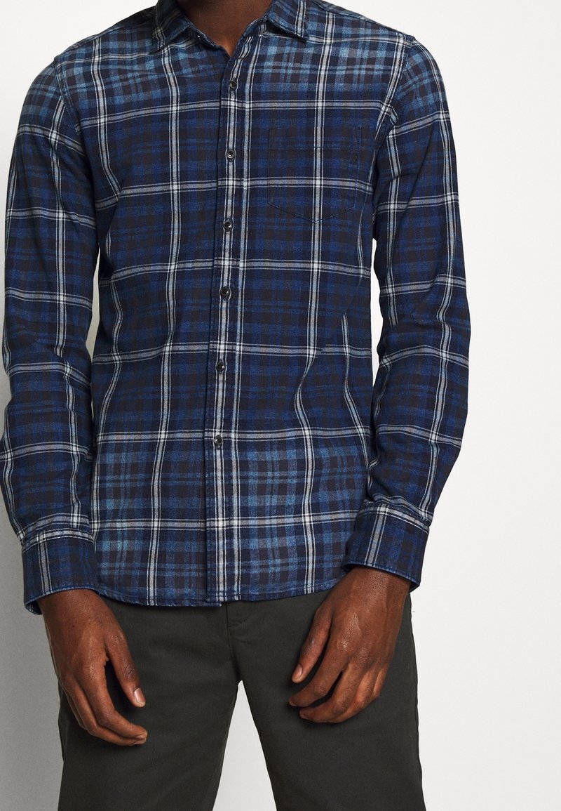 Uomo che indossa una camicia a quadri blu e nera con maniche lunghe e pantaloni scuri, in piedi con le mani rilassate ai lati.