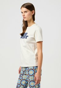 Camisa de algodón crema con mangas cortas que presenta el texto "GO WEST" en azul marino. Combinada con pantalones azules estampados con diseños florales.
