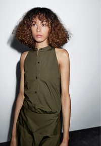 Olijfgroen mouwloze top met een hoge kraag, met een doorlopende knoopsluiting aan de voorkant en een gathered taille voor een ontspannen pasvorm. Soepele stoftextuur.