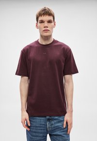 Bordo bombažna majica s krožnim izrezom in kratkimi rokavi. Na sprednji strani majhen logotip "HUGO". Nošena z modrimi jeansa.