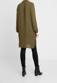 Tunic top verde oliva a maniche lunghe con colletto alto, tessuto testurizzato, fessure laterali, abbinato a leggings neri e stivaletti.