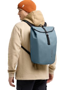 Sac à dos imperméable bleu clair avec fermeture enroulable, sangle noire et logo, porté sur une veste en polaire beige.