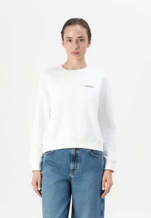 Felpa bianca con polsini e orlo a coste, caratterizzata da un piccolo logo "Guess Jeans" sul petto. Abbinata a jeans azzurri chiari.