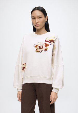 Frau trägt einen weißen Pullover mit farbenfrohen abstrakten Blumendesigns und braune Hosen, steht vor einem schlichten hellen Hintergrund.