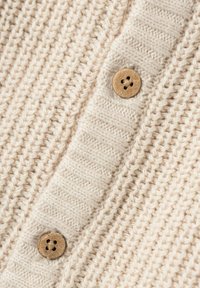 Beige gestrickte Strickjacke mit vertikalen und horizontalen Rippenmustern, ausgestattet mit zwei Holzknöpfen mit jeweils vier Löchern.