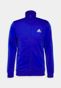 Veste de survêtement zippée bleue Adidas avec col montant et logo blanc Adidas sur la poitrine gauche, manches longues et poignets et bas côtelés.