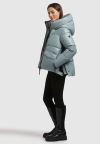 Chaqueta acolchada y de gran tamaño en verde menta, con un cuello alto, cierre de cremallera frontal y dos bolsillos delanteros; combinada con pantalones negros ajustados y botas altas negras.