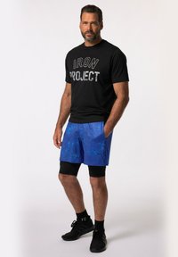 T-shirt noir avec "IRON PROJECT" en gris métallique, associé à un short bleu avec un motif abstrait texturé. Des chaussures de sport et des chaussettes complètent le look.