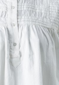 Balta kokvilnas blūze ar krokotu tekstūru, priekšējiem pogām un savilktām jostām. Iespējams vertikāls šuvums un smalka spīdība.