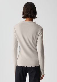 Helles beige Langarmoberteil aus weichem Material mit einem gewellten Saum und Ausschnitt. Die Rückseite ist enganliegend und hat eine glatte Textur.