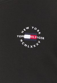 Tissu noir avec logo Tommy Hilfiger brodé, texte blanc « NEW YORK MCMLXXV » entourant un drapeau rectangulaire rouge, blanc et bleu marine.