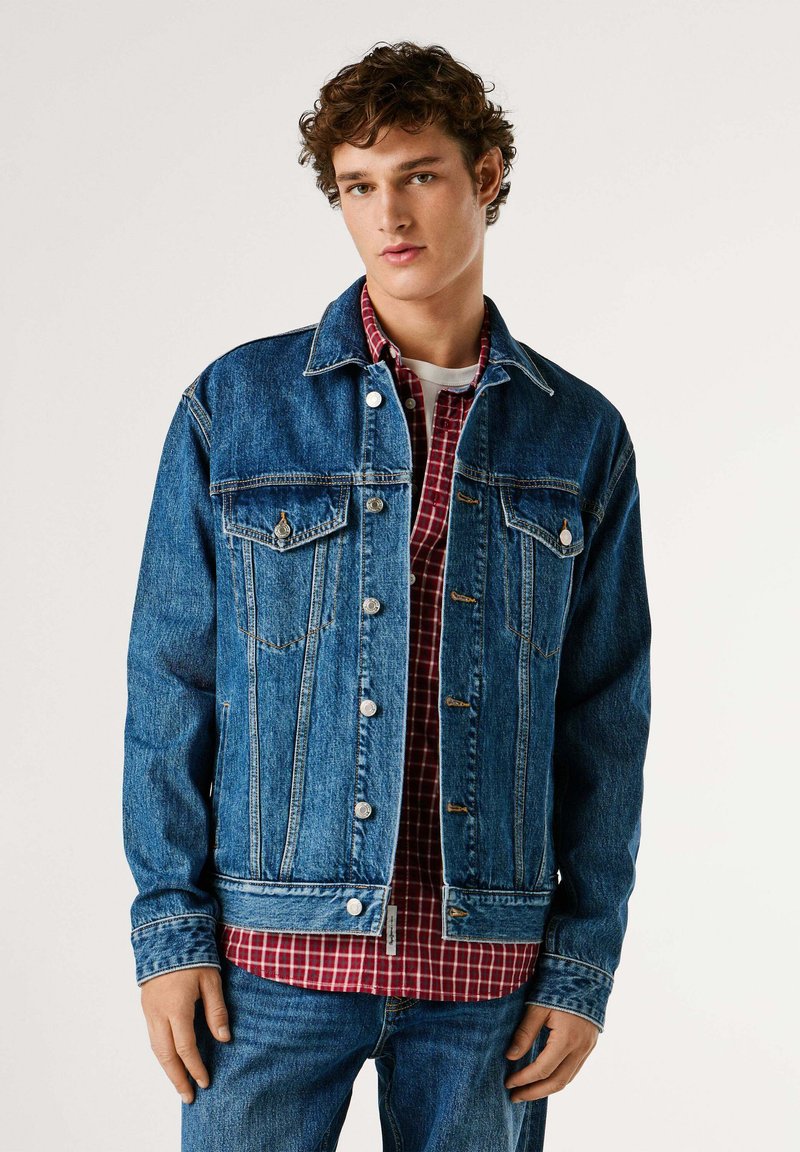 Chaqueta vaquera en azul oscuro, con herrajes plateados, dos bolsillos en el pecho y detalles cosidos. Llevada sobre una camisa a cuadros roja.