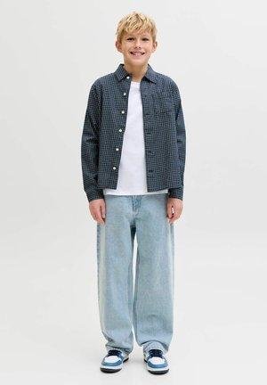 Pārbaudīta tumši zila pogām aizpogājama krekls pār baltu T-kreklu, kombinēts ar gaiši ziliem vaļīgiem džinsiem un ziliem akcentiem apavos.