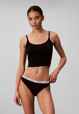 Femme portant un débardeur noir court et des sous-vêtements Calvin Klein noirs avec une ceinture à logo blanche, posant devant un fond gris uni.