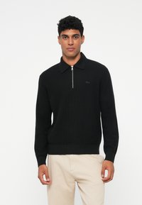 Schwarzer Strick-Zipfelpullover mit spitzem Kragen, der ein dezentes Logo und gerippte Bündchen aufweist, kombiniert mit hellen Hosen.