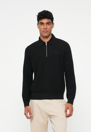 Maglione zip-up nero in maglia con colletto a punta, caratterizzato da un logo discreto e polsini a coste, abbinato a pantaloni chiari.