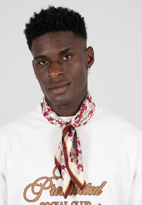 Silket skjerf med blomstermønster i beige, rosa og rødt, knyttet rundt nakken, med en kontrasterende kant i mørkerød og gul.