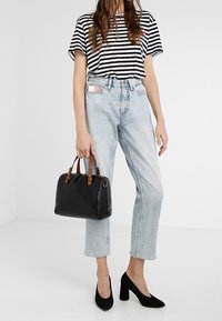 Sort håndtaske med brune læderdetaljer, blød tekstur, rektangulær form. Båret med stribet skjorte og lyseblå højtaljede jeans.