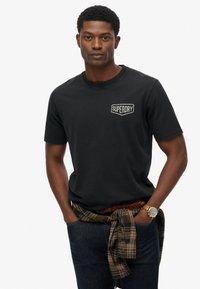 Camiseta de algodón negra con un pequeño logo bordado de 'Superdry' en el pecho. El modelo la combina con jeans oscuros y una camisa de cuadros atada a la cintura.