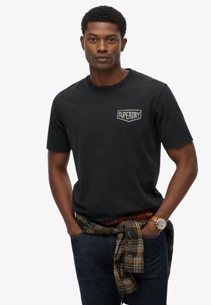 T-shirt en coton noir avec un petit logo brodé 'Superdry' sur la poitrine. Le modèle l'associe à un jean noir et une chemise à carreaux nouée à la taille.
