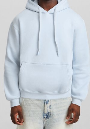 Person trägt ein hellblaues Kapuzen-Sweatshirt mit Vordertasche und Kordelzug, kombiniert mit hellblauen Jeans.