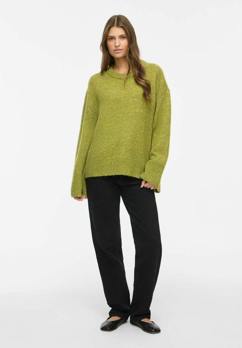 Femme aux longs cheveux bruns portant un pull vert oversize, un pantalon noir et des ballerines noires, debout devant un fond uni clair.