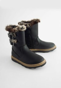 Schwarze Knöchelstiefel mit Kunstfellbesatz, doppelten Riemenakzenten und einer strukturierten Sohle. Hergestellt aus glattem, lederähnlichem Material. Reißverschluss an der Seite.