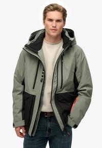 Ung mand iført en lys grøn og sort hættejakke med rød lynlåsdetalje over en beige sweater og blå jeans.