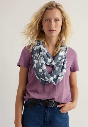 Noor naine laineliste blondide juustega, kandes lilla lühikeste varrukatega särki, sinist lillelist sall, siniseid teksaseid ja musta vööd, seisab käsi taskus.