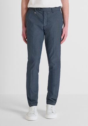 Pantalones azul marino con tejido texturizado, diseño ajustado, trabillas para cinturón y cierre frontal. Combinados con zapatillas blancas.