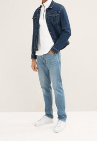 Chaqueta de mezclilla en azul oscuro, polo blanco con cuello con cremallera, jeans de mezclilla azul claro y zapatillas blancas. Atuendo casual en capas con cortes clásicos.