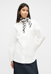 Γυναίκα που φορά ένα λευκό πουκάμισο με κουμπιά, σκούρα τζιν φούστα και μαύρο-άσπρο ριγωτό φουλάρι στο λαιμό, στέκεται μπροστά σε απλό φόντο.