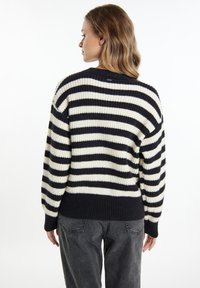 Frau in einem schwarz-weißen, horizontal gestreiften Pullover und dunklen Jeans, die mit dem Rücken zu einem schlichten weißen Hintergrund steht.