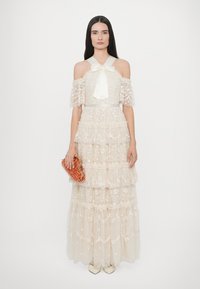 IVORELLAGEN JURK MET OFF-SHOULDER MOUWEN, BLOEMENBORDUURSEL EN EEN SATIJNEN STRIK BIJ DE KRAAG; SAMENGESTELD MET EEN GEVEKT EN ORANJE HANDBAG.