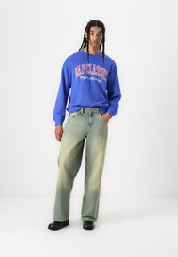 Sudadera azul con "GAP CLASSIC" en texto blanco, combinada con jeans de pierna ancha verde claro. El modelo lleva zapatos negros, de pie sobre un fondo blanco.