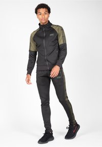 Chándal deportivo negro con acentos de hombro en verde oliva, con bolsillos con cremallera y cinta de marca a lo largo de los pantalones. Textura suave, corte ajustado.