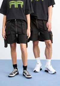 Calças de carga pretas com zíperes, bolsos laterais e costuras contrastantes. Usadas com t-shirts oversized e sapatilhas, contra um fundo claro.