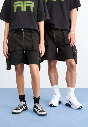 Schwarze Cargo-Shorts mit Reißverschlüssen, Seitentaschen und kontrastierendem Nähen. Getragen mit übergroßen T-Shirts und Sportschuhen, vor einem hellen Hintergrund.