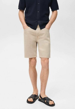 Beige denim shorts with a straight cut and two front pockets, paired with a navy button-up shirt and black cross-strap sandals.