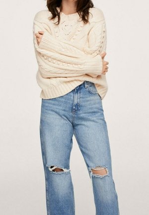 Kobieta ubrana w kremowy sweter z grubym splotem i długimi rękawami oraz jasnoniebieskie, postrzępione dżinsy, z założonymi na piersi skrzyżowanymi ramionami.
