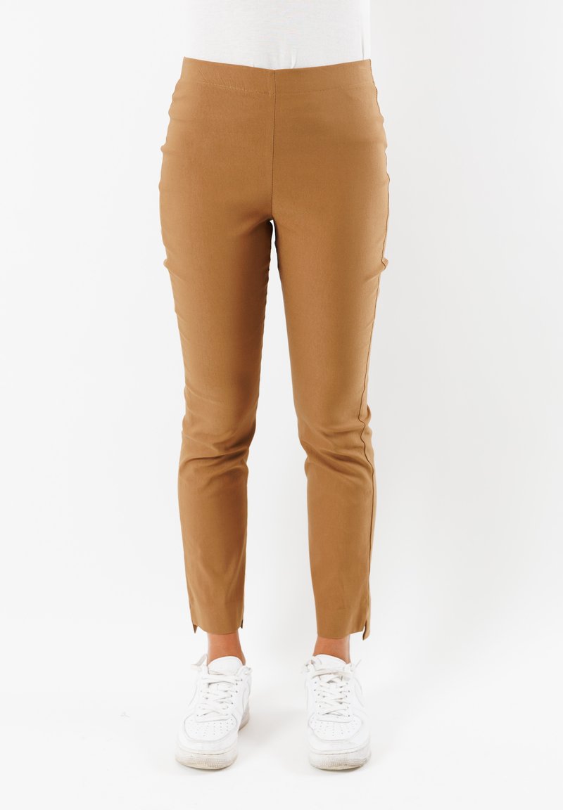 Pantaloni marroni aderenti con una texture liscia, caratterizzati da un'alta vita e un dettaglio a fessura alla caviglia, abbinati a sneakers bianche.