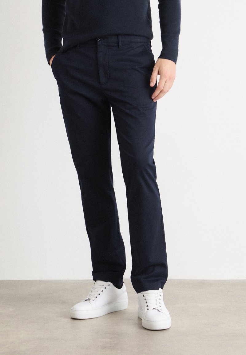 Pantaloni in cotone blu navy con taglio dritto, dotati di vita piatta, tasche e passanti per la cintura classici, abbinati a sneaker bianche.