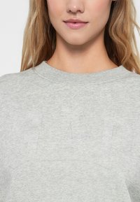 Světle šedá mikina s kulatým výstřihem, která má na přední straně jemný ražený design. Měkká, texturovaná tkanina viditelná zblízka.