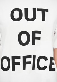 Λευκό βαμβακερό μπλουζάκι με μεγάλα έντονα μαύρα γράμματα που γράφουν «ΕΚΤΟΣ ΓΡΑΦΕΙΟΥ» στο κέντρο του μπροστινού μέρους. Κοντά μανίκια και στρογγυλή λαιμόκοψη.